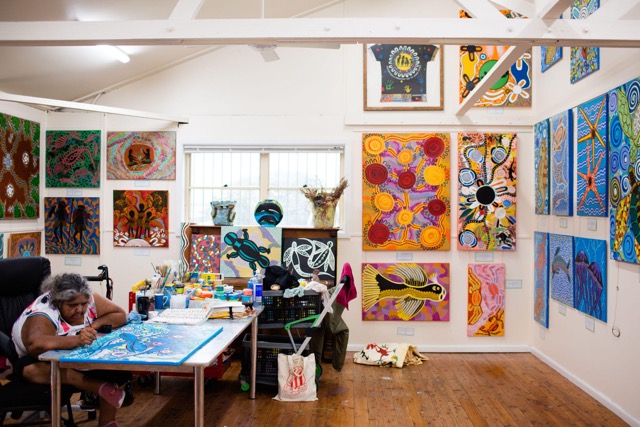 Artist painting at a table in the Coomaditchie studio, surrounded by vibrant Aboriginal artworks