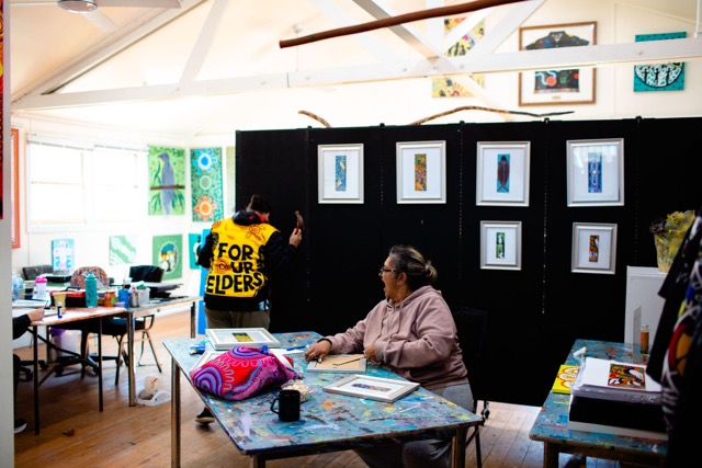 Coomaditchie studio with framed artworks and an artist in a For Our Elders jacket hanging a display