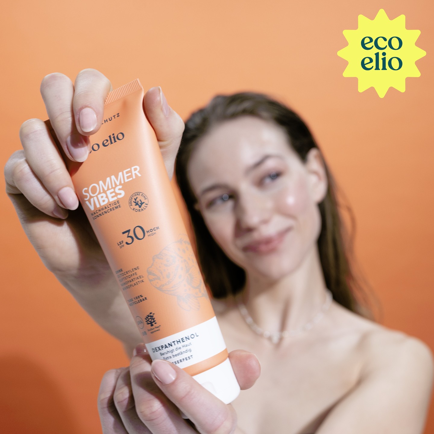 Model holding an orange eco elio Sommer Vibes SPF30 sunscreen tube against an orange background