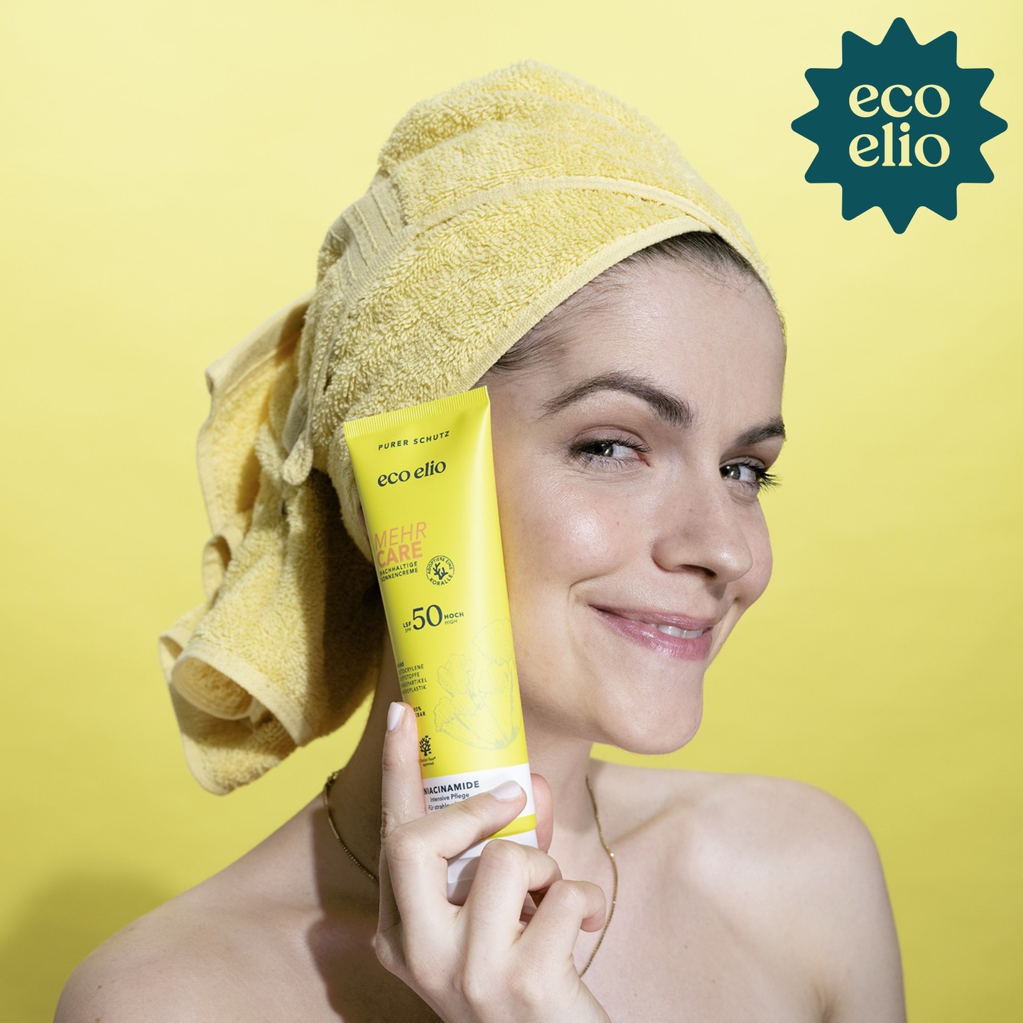 Model in a yellow towel turban holding a yellow eco elio Mehr Care SPF50 sunscreen tube against a yellow background